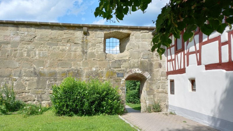 Historische Stadtmauer mit kleinem Torbogen und leicht nach links versetztem "Fenster", rechts an die Mauer anschlie&szlig;end ein Fachwerkhaus mit roten Holzbalken. Im Vordergrund eine Wiese mit Str&auml;uchern., &copy; Petra Natzkowski