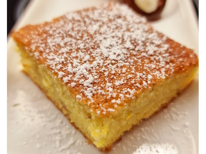 Ein saftiges Stück Zitronenkuchen, bestreut mit Puderzucker, liegt auf einem Teller. Im Hintergrund ist ein Klecks Sahne zu sehen., © Kultweine-Kochwelten-Kunstücke - Kochschule Sonja Lenz Ein saftiges Stück Zitronenkuchen, bestreut mit Puderzucker, liegt auf einem Teller. Im Hintergrund ist ein Klecks Sahne zu sehen., © Kultweine-Kochwelten-Kunstücke - Kochschule Sonja Lenz