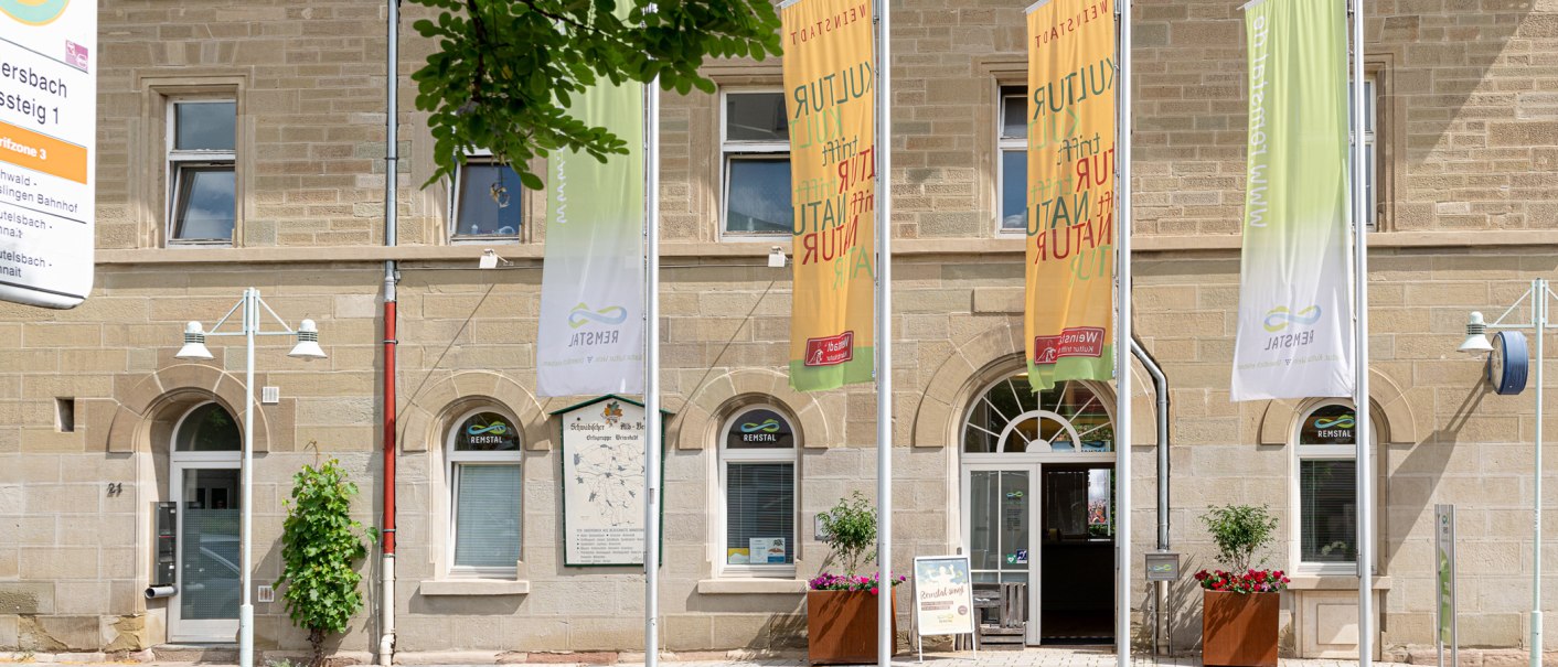 Außenansicht eines Tourismusbüros mit mehreren Fahnen und Blumenkästen. Das Gebäude hat eine sandfarbene Fassade und eine Uhr an der Wand., © Remstal Tourismus, Peter Schuster