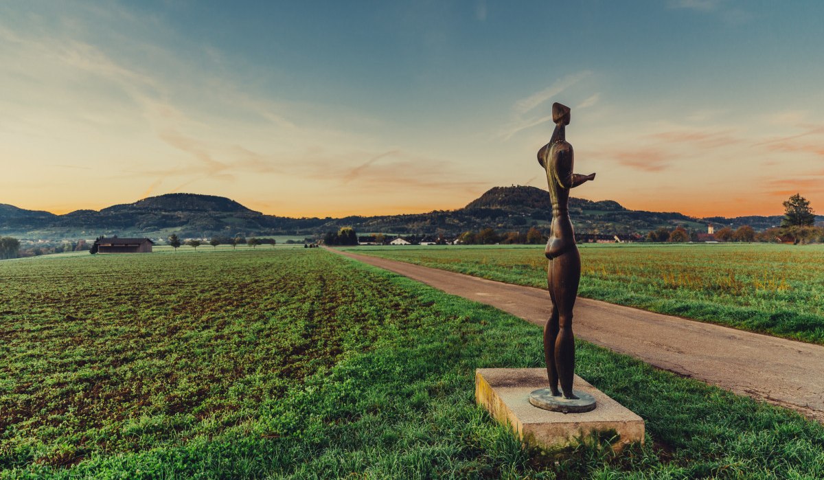 Eine Skulptur steht auf einem Feldweg, umgeben von grünen Feldern und Hügeln im Hintergrund, während die Sonne untergeht., © Landratsamt Ostalbkreis