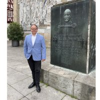Ein Mann in einem blauen Sakko steht neben einem Denkmal von Gottlieb Daimler in Schorndorf. Im Hintergrund ist ein Wandrelief zu sehen., &copy; Eigenbetrieb Tourismus und Citymanagement Schorndorf