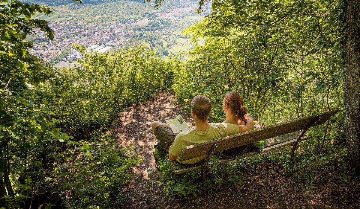 Ein Paar sitzt auf einer Bank im Wald und blickt auf das Lenninger Tal. Die Landschaft ist grün und idyllisch., © hochgehberge Ein Paar sitzt auf einer Bank im Wald und blickt auf das Lenninger Tal. Die Landschaft ist grün und idyllisch., © hochgehberge