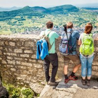 Drei Wanderer mit Rucksäcken stehen auf einer Mauer und genießen den Ausblick auf die grüne Landschaft von Hohenneuffen., © hochgehberge Drei Wanderer mit Rucksäcken stehen auf einer Mauer und genießen den Ausblick auf die grüne Landschaft von Hohenneuffen., © hochgehberge