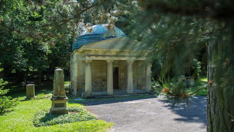 Ein Mausoleum mit Säulen steht in einem grünen Park, umgeben von Bäumen und einem Grabstein. Die Szene wirkt friedlich und historisch., © Copyright: Tourismus & Events Ludwigsburg, Fotograf: Benjamin Stollenberg Ein Mausoleum mit Säulen steht in einem grünen Park, umgeben von Bäumen und einem Grabstein. Die Szene wirkt friedlich und historisch., © Copyright: Tourismus & Events Ludwigsburg, Fotograf: Benjamin Stollenberg