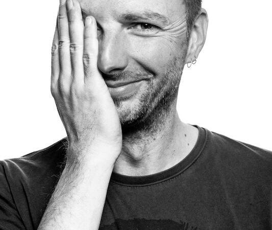 Ein Mann lächelt in die Kamera und hält eine Hand vor sein Gesicht. Er trägt ein T-Shirt und hat einen Ohrring. Schwarz-weiß Fotografie., © Kulturhaus Schwanen