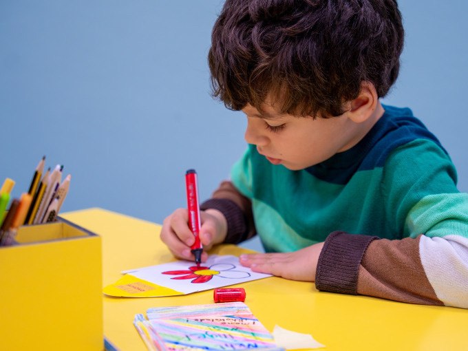 Ein Kind malt konzentriert mit einem roten Stift auf einem Blatt Papier an einem gelben Tisch. Im Hintergrund sind Buntstifte zu sehen., &copy; Alfred Ritter GmbH &amp; Co. KG | Waldenbuch
