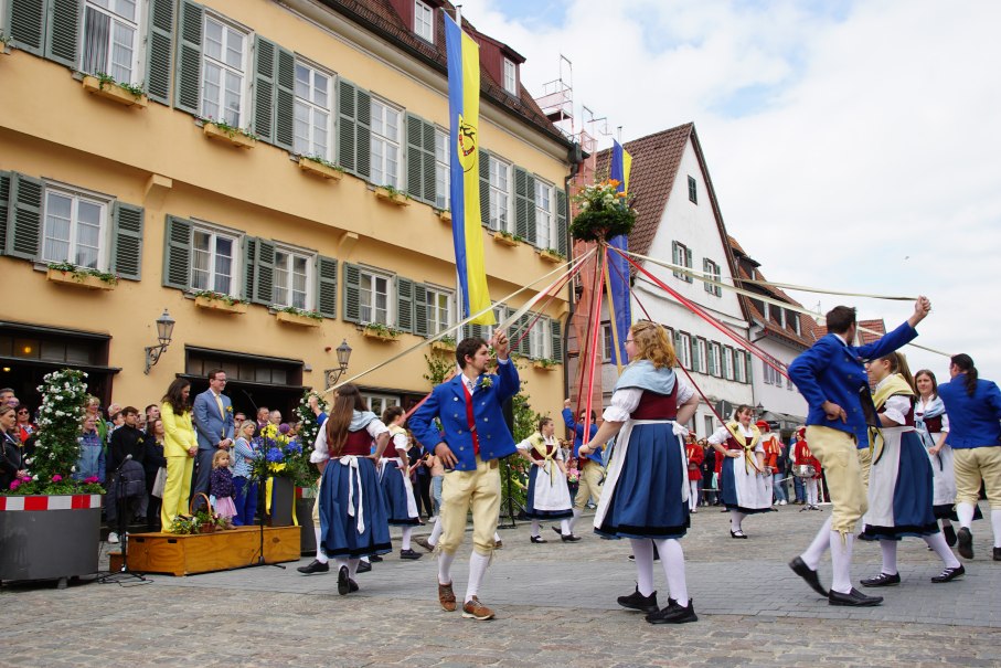 Tänzer in traditionellen Trachten führen einen Bändertanz um einen Maibaum vor einem Rathaus auf. Zuschauer beobachten das Spektakel., © T. Krytzner