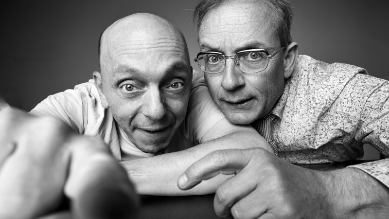 Zwei M&auml;nner in Schwarz-Wei&szlig;-Nahaufnahme, einer zeigt auf die Kamera, der andere schaut neugierig. Beide wirken humorvoll und interessiert.