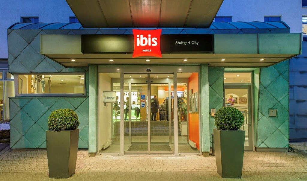 Eingang des Ibis Hotels Stuttgart City mit beleuchtetem Schild und Glasdrehtür, flankiert von zwei Pflanzen in modernen Pflanzgefäßen., © TOMAS