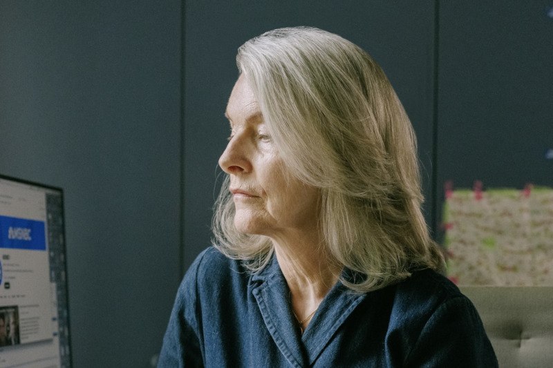 Eine Frau mit grauem Haar sitzt nachdenklich in einem Raum, schaut zur Seite. Im Hintergrund sind ein Bildschirm und eine Pinnwand zu sehen., © Kulturamt der Stadt Esslingen am Neckar