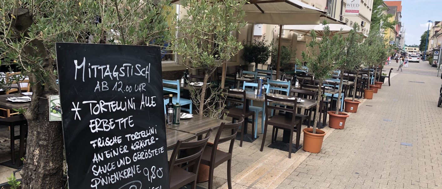 Ein Straßencafé mit Tischen und Stühlen unter Sonnenschirmen. Eine Menütafel bietet Tortellini und Fleischgerichte an. Olivenbäume in Töpfen säumen den Weg., © Antipasto Wein und mehr