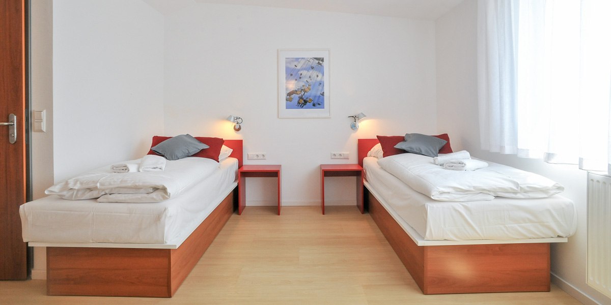 Helles Zweibettzimmer mit zwei Einzelbetten, roten Nachttischen und Wandlampen. Ein Bild hängt an der weißen Wand, Fenster mit weißen Vorhängen., © Gästehaus ABZ Stuttgart Helles Zweibettzimmer mit zwei Einzelbetten, roten Nachttischen und Wandlampen. Ein Bild hängt an der weißen Wand, Fenster mit weißen Vorhängen., © Gästehaus ABZ Stuttgart