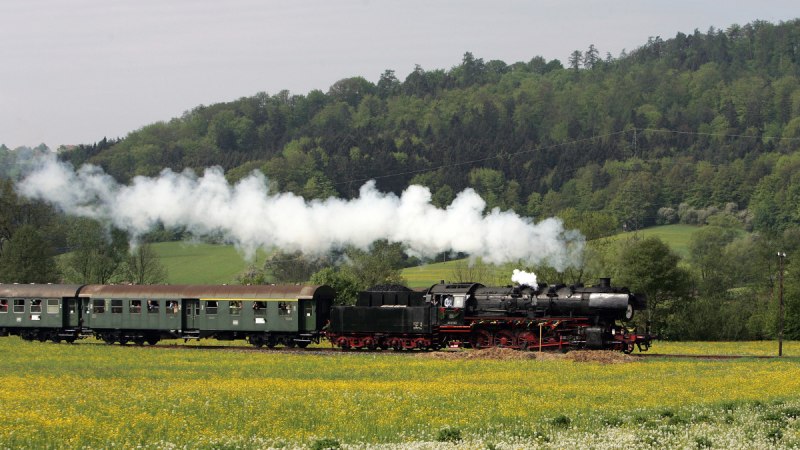 Eine Dampflok zieht gr&uuml;ne Waggons durch eine bl&uuml;hende Wiese, umgeben von bewaldeten H&uuml;geln. Rauch steigt aus der Lokomotive auf., &copy; E-Bike-Region Stuttgart