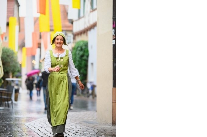 Frau in grüner historischer Kleidung geht lächelnd durch eine gepflasterte Straße mit bunten Fahnen im Hintergrund., © Eigenbetrieb Tourismus und Citymanagement Schorndorf Frau in grüner historischer Kleidung geht lächelnd durch eine gepflasterte Straße mit bunten Fahnen im Hintergrund., © Eigenbetrieb Tourismus und Citymanagement Schorndorf
