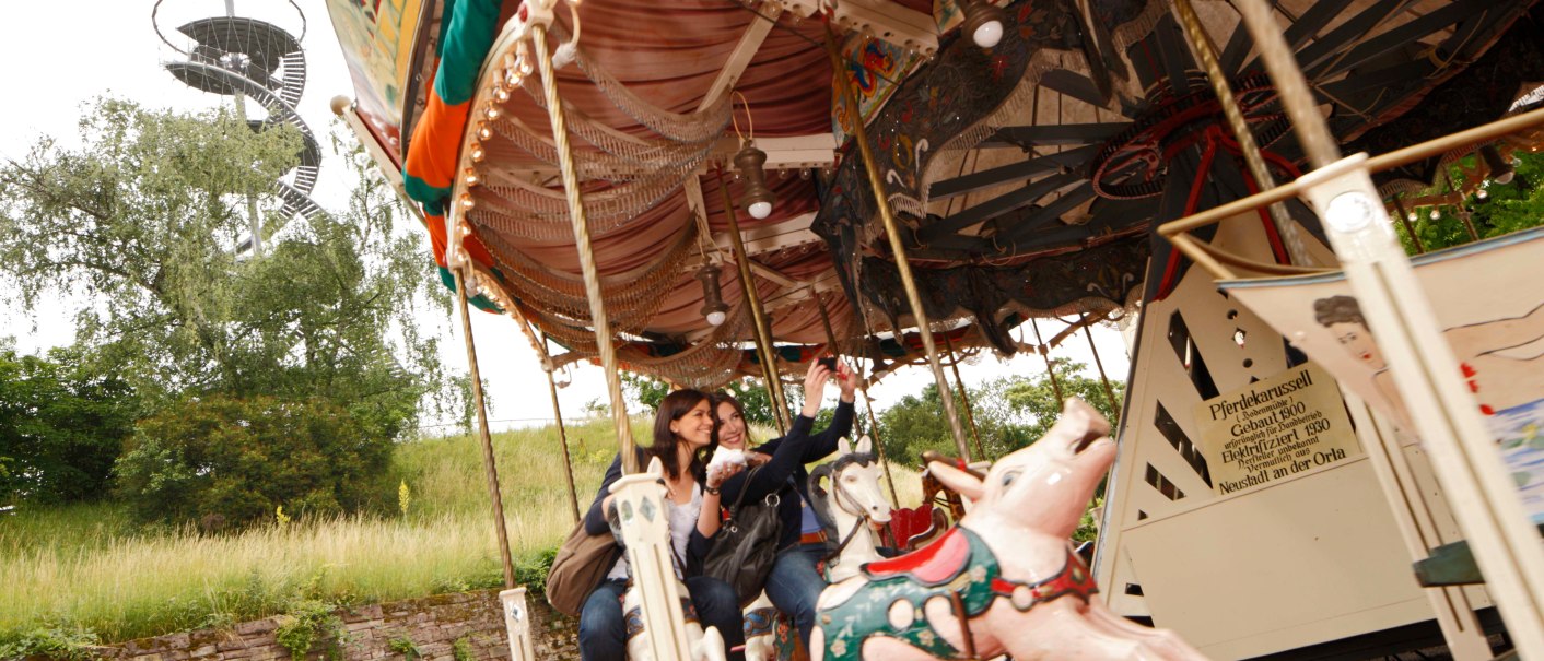 Zwei Personen fahren auf einem nostalgischen Karussell mit Pferdefiguren. Im Hintergrund ist ein spiralförmiger Aussichtsturm zu sehen., © SMG, Christoph Düpper Zwei Personen fahren auf einem nostalgischen Karussell mit Pferdefiguren. Im Hintergrund ist ein spiralförmiger Aussichtsturm zu sehen., © SMG, Christoph Düpper