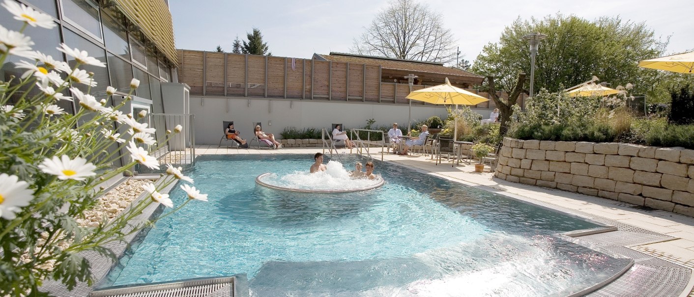 Außenbereich der Barbarossa-Thermen mit Pool, sprudelndem Wasser und Liegestühlen. Menschen entspannen unter Sonnenschirmen. Blumen und Bäume im Hintergrund., © Barbarossa-Thermen Göppingen