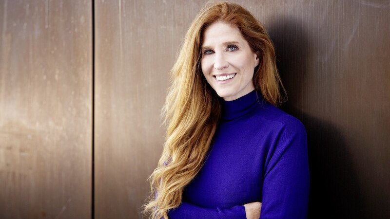 Eine Frau mit langen roten Haaren tr&auml;gt einen blauen Pullover und lehnt l&auml;chelnd an einer metallischen Wand., &copy; Osiander Vertriebsgesellschaft mbH & Co. KG