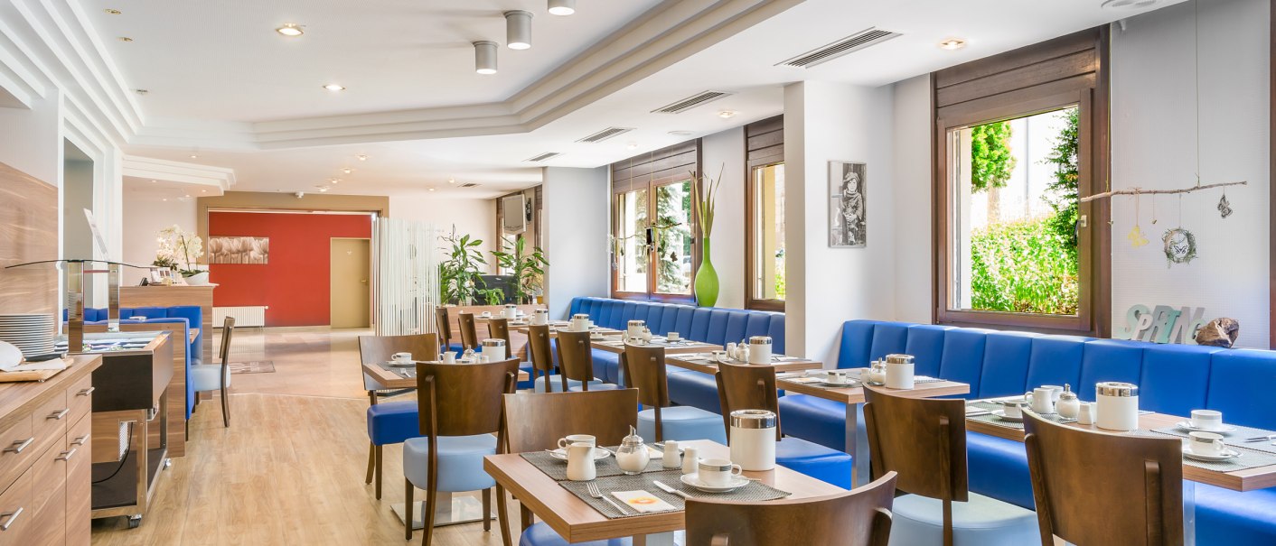 Heller Frühstücksraum mit blauen Sitzbänken, Holztischen und -stühlen. Große Fenster lassen viel Licht herein, Pflanzen dekorieren den Raum., © TOMAS