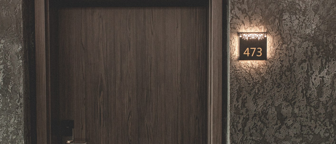 Eine Holztür mit der Zimmernummer 473 an einer strukturierten Wand. Die Nummer ist beleuchtet., © Plaza Residence Stuttgart-Ditzingen Eine Holztür mit der Zimmernummer 473 an einer strukturierten Wand. Die Nummer ist beleuchtet., © Plaza Residence Stuttgart-Ditzingen