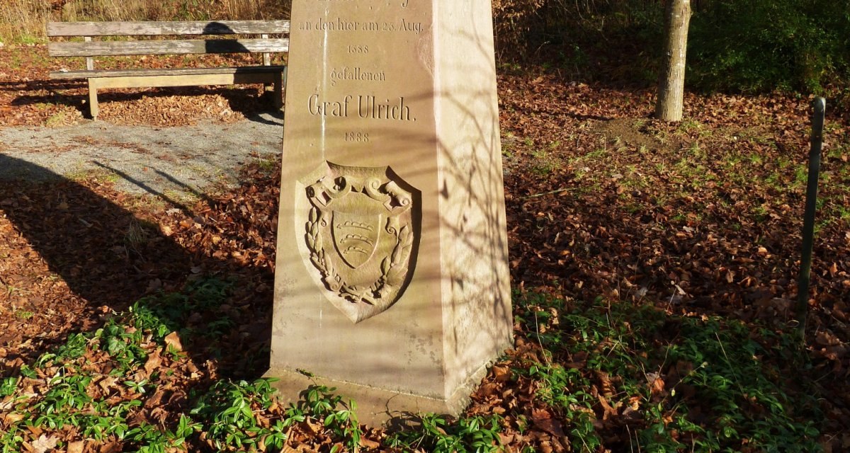 Ein Gedenkstein im Wald erinnert an Graf Ulrich. Der Stein steht auf Laub und ist von Pflanzen umgeben. Eine Bank ist im Hintergrund sichtbar., © Natur.Nah. Schönbuch & Heckengäu Ein Gedenkstein im Wald erinnert an Graf Ulrich. Der Stein steht auf Laub und ist von Pflanzen umgeben. Eine Bank ist im Hintergrund sichtbar., © Natur.Nah. Schönbuch & Heckengäu