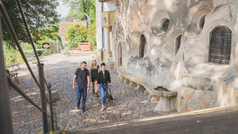 Vier Personen spazieren auf einem gepflasterten Weg neben einer historischen Steinmauer in Schw&auml;bisch Gm&uuml;nd. Im Hintergrund sind B&auml;ume und ein kleines Geb&auml;ude zu sehen.