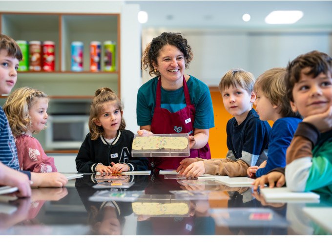 Eine Frau in Schürze zeigt Kindern eine Schokoladentafel in einer modernen Küche. Die Kinder schauen interessiert zu., © Alfred Ritter GmbH & Co. KG | Waldenbuch Eine Frau in Schürze zeigt Kindern eine Schokoladentafel in einer modernen Küche. Die Kinder schauen interessiert zu., © Alfred Ritter GmbH & Co. KG | Waldenbuch
