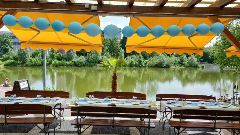 Gedeckte Tische unter gelben Markisen, dekoriert mit blauen Luftballons, mit Blick auf einen See und grüne Bäume im Hintergrund., © Gaststätte Goldbachsee Gedeckte Tische unter gelben Markisen, dekoriert mit blauen Luftballons, mit Blick auf einen See und grüne Bäume im Hintergrund., © Gaststätte Goldbachsee