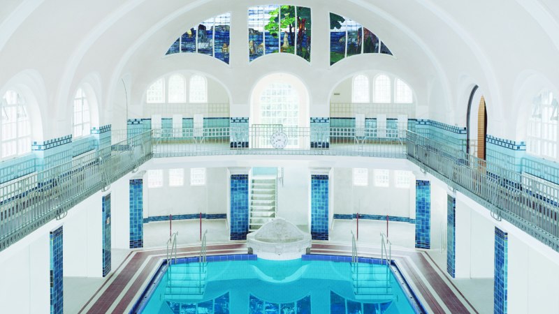 Innenansicht des Merkel'schen Schwimmbads in Esslingen. Das Becken ist von blauen Fliesen umgeben, mit hohen Fenstern und einem kunstvollen Glasfenster., © Esslinger Stadtmarketing & Tourismus GmbH