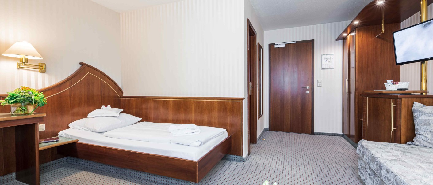 Ein gemütliches Hotelzimmer mit Einzelbett, Holzmöbeln, einem kleinen Tisch mit Blumen und einem Fernseher an der Wand., © TOMAS