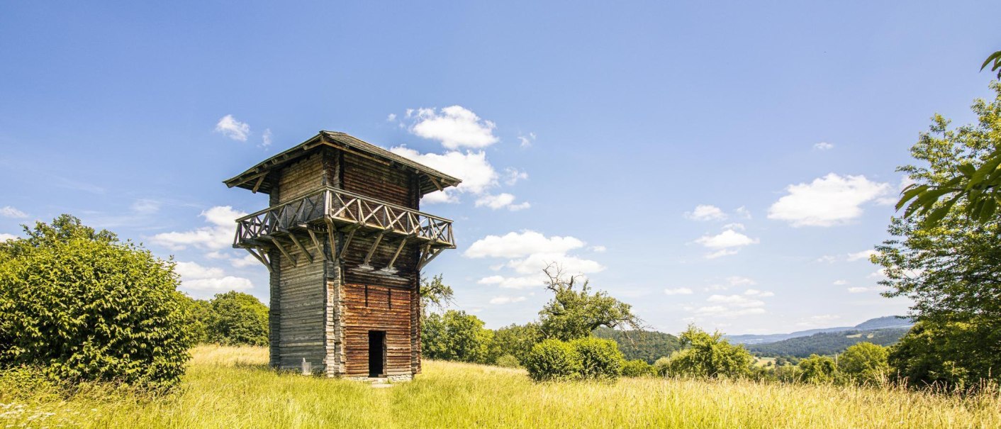 Ein hölzerner Wachturm auf einer grünen Wiese unter klarem Himmel. Der Turm ist von Bäumen umgeben und die Landschaft erstreckt sich im Hintergrund., © SMG, Sarah Schmid Ein hölzerner Wachturm auf einer grünen Wiese unter klarem Himmel. Der Turm ist von Bäumen umgeben und die Landschaft erstreckt sich im Hintergrund., © SMG, Sarah Schmid