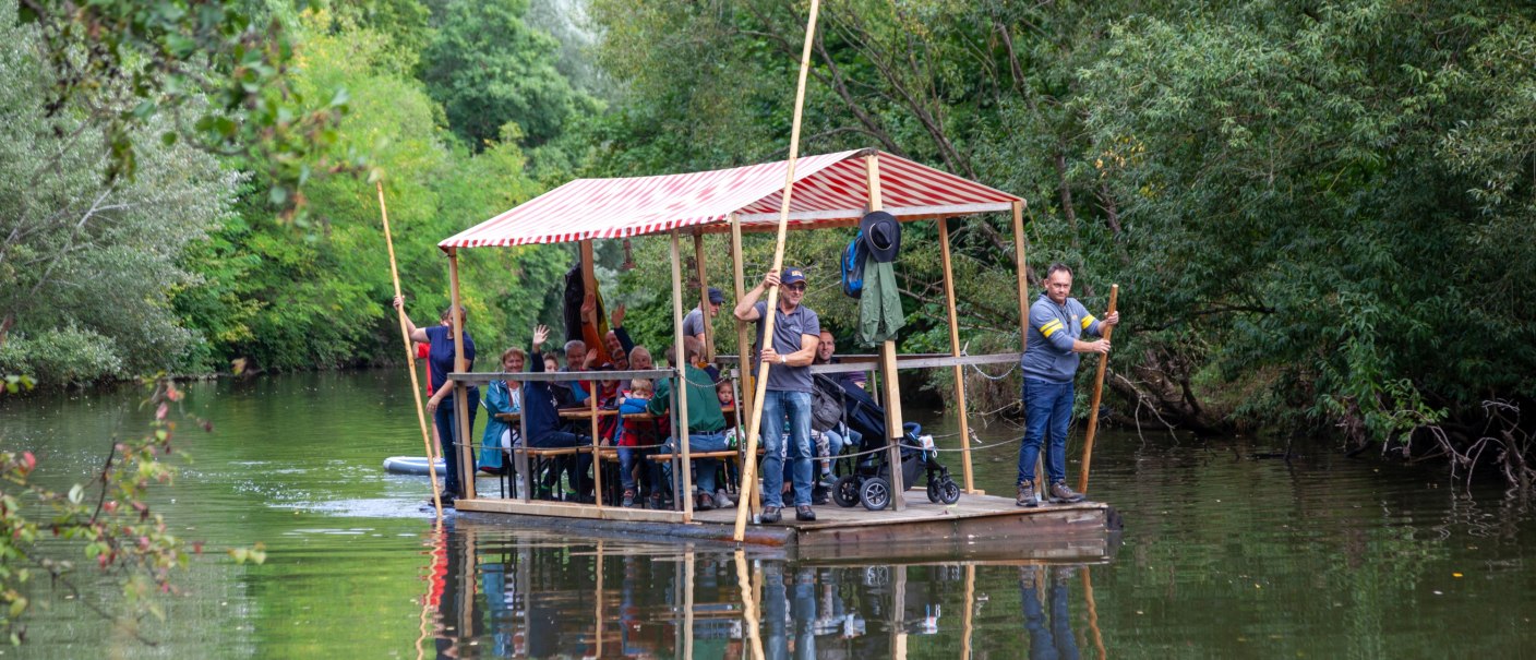 überdachtes Holzfloß mit Menschen auf dem Fluß, an den Ufern Büsche und Bäume, © Stadt Gaildorf