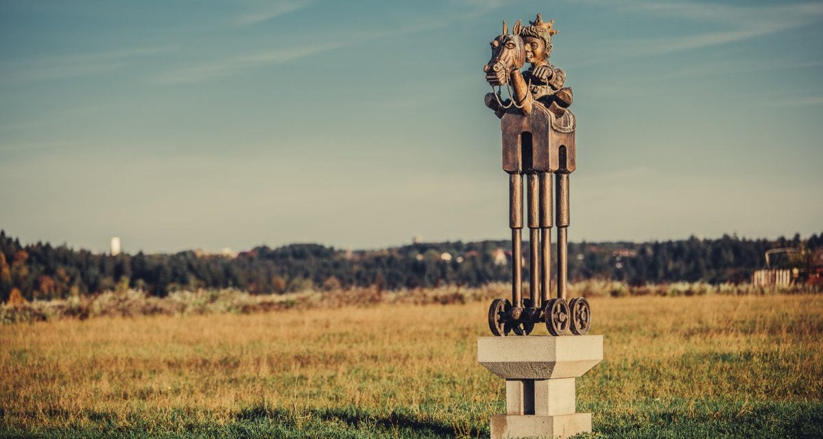 Eine Skulptur zeigt einen Reiter auf einem Pferd mit Rädern, platziert auf einem Sockel in einer weiten Wiesenlandschaft unter blauem Himmel., © Landratsamt Ostalbkreis Eine Skulptur zeigt einen Reiter auf einem Pferd mit Rädern, platziert auf einem Sockel in einer weiten Wiesenlandschaft unter blauem Himmel., © Landratsamt Ostalbkreis