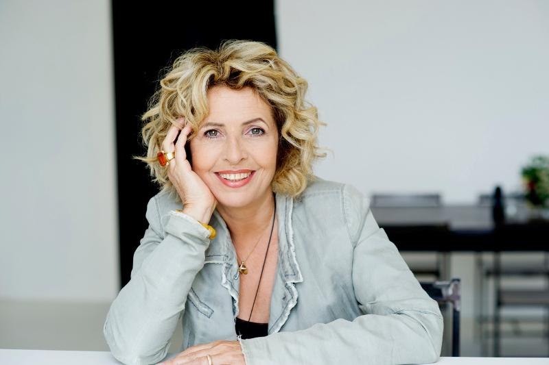 Frau mit lockigem Haar lächelt, stützt den Kopf auf die Hand. Sie trägt eine helle Jacke und sitzt an einem Tisch., © Backnang