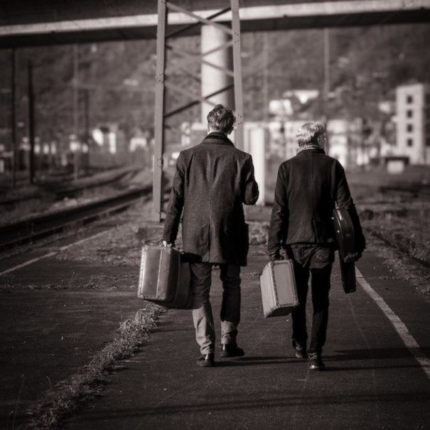 Zwei Personen gehen mit Koffern auf einem leeren Bahnsteig. Die Szene ist in Schwarz-Weiß gehalten, was eine nostalgische Stimmung erzeugt., © richtungdichtung Zwei Personen gehen mit Koffern auf einem leeren Bahnsteig. Die Szene ist in Schwarz-Weiß gehalten, was eine nostalgische Stimmung erzeugt., © richtungdichtung