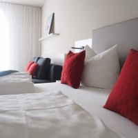 Modernes Hotelzimmer mit weißen Bettlaken, roten Kissen und einem Sofa. Ein Bild hängt an der Wand, helle Vorhänge lassen Licht herein., © TOMAS Modernes Hotelzimmer mit weißen Bettlaken, roten Kissen und einem Sofa. Ein Bild hängt an der Wand, helle Vorhänge lassen Licht herein., © TOMAS