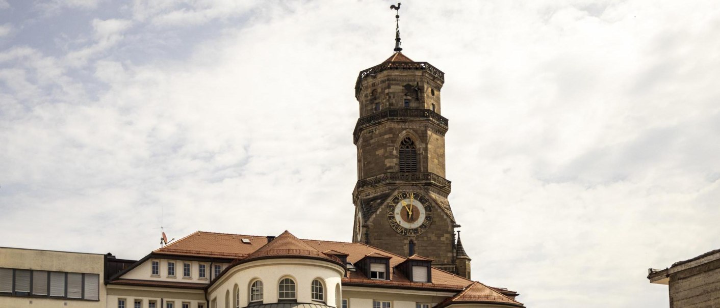 Turm der Stiftskirche Stuttgart mit Uhr und Wetterhahn, umgeben von Gebäuden unter bewölktem Himmel., © Stuttgart Marketing GmbH, Sarah Schmid Turm der Stiftskirche Stuttgart mit Uhr und Wetterhahn, umgeben von Gebäuden unter bewölktem Himmel., © Stuttgart Marketing GmbH, Sarah Schmid