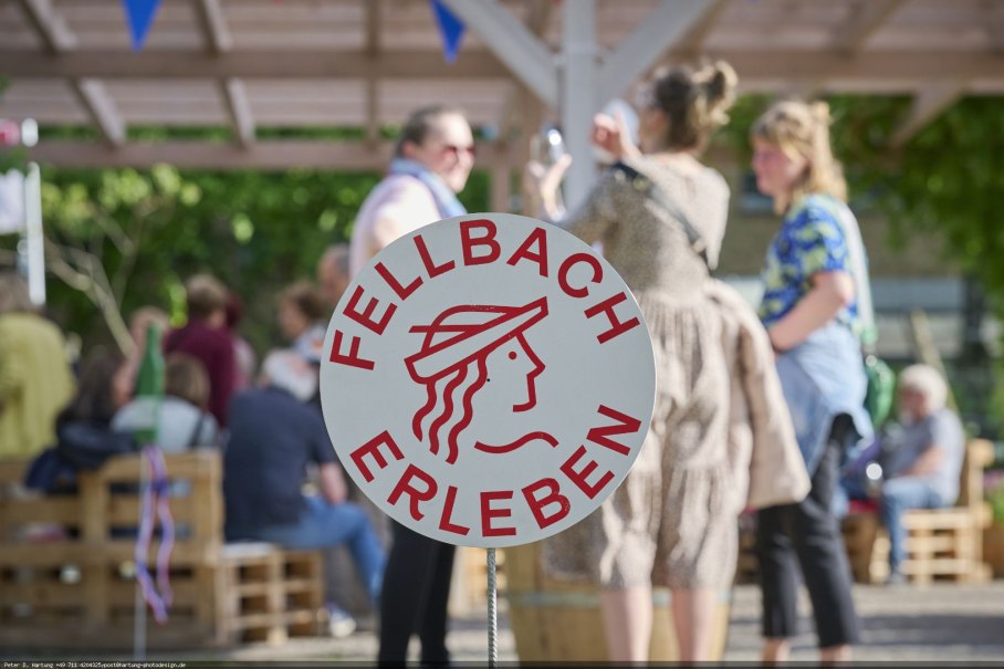Ein Schild mit der Aufschrift 'Fellbach Erleben' steht im Vordergrund. Im Hintergrund sind Menschen bei einer Outdoor-Veranstaltung zu sehen., &copy; Stadt Fellbach