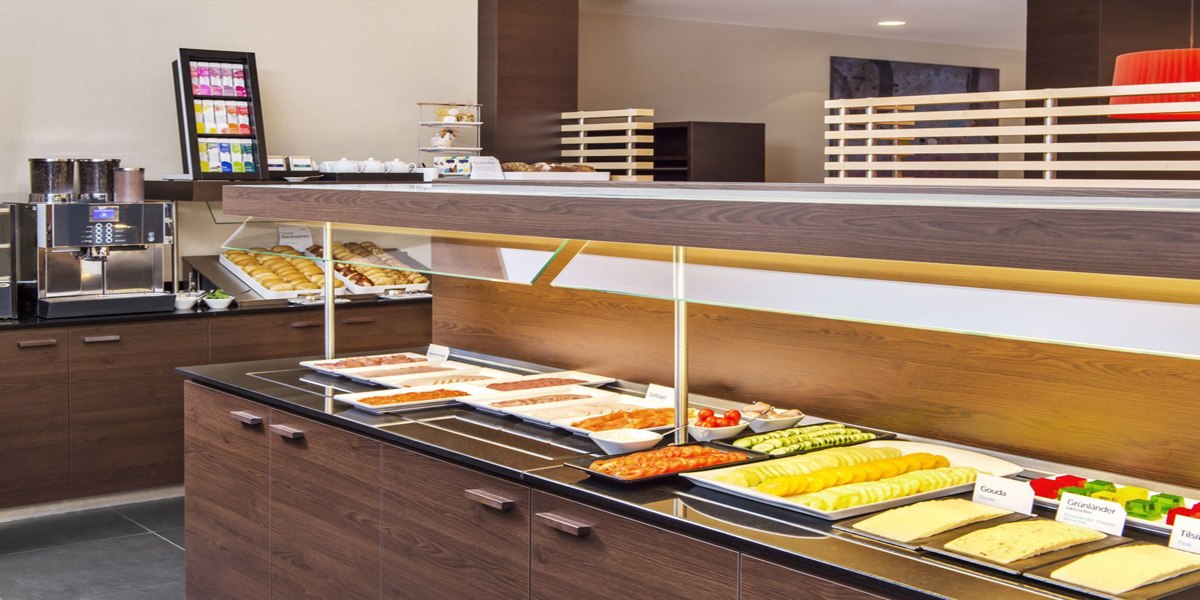Ein Frühstücksbuffet mit verschiedenen Aufschnitten, Käse, Gebäck und einer Kaffeemaschine in einem modernen, hellen Raum., © NH Ludwigsburg
