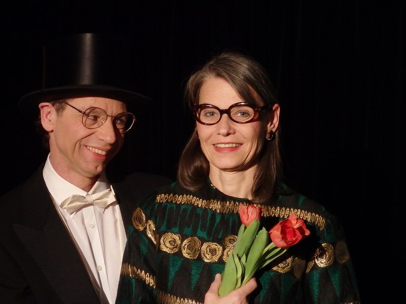 Ein Paar in eleganter Kleidung, der Mann mit Zylinder, die Frau mit Blumenstrau&szlig;. Beide l&auml;cheln und tragen Brillen., &copy; Galli Theater Backnang