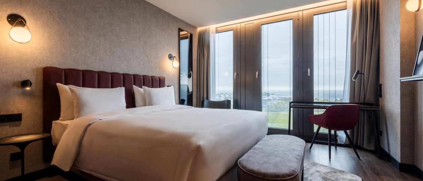 Modernes Hotelzimmer mit großem Bett, Schreibtisch und Blick auf Weinberge durch große Fenster. Elegante Beleuchtung und stilvolle Einrichtung., © Radisson Hotel Group