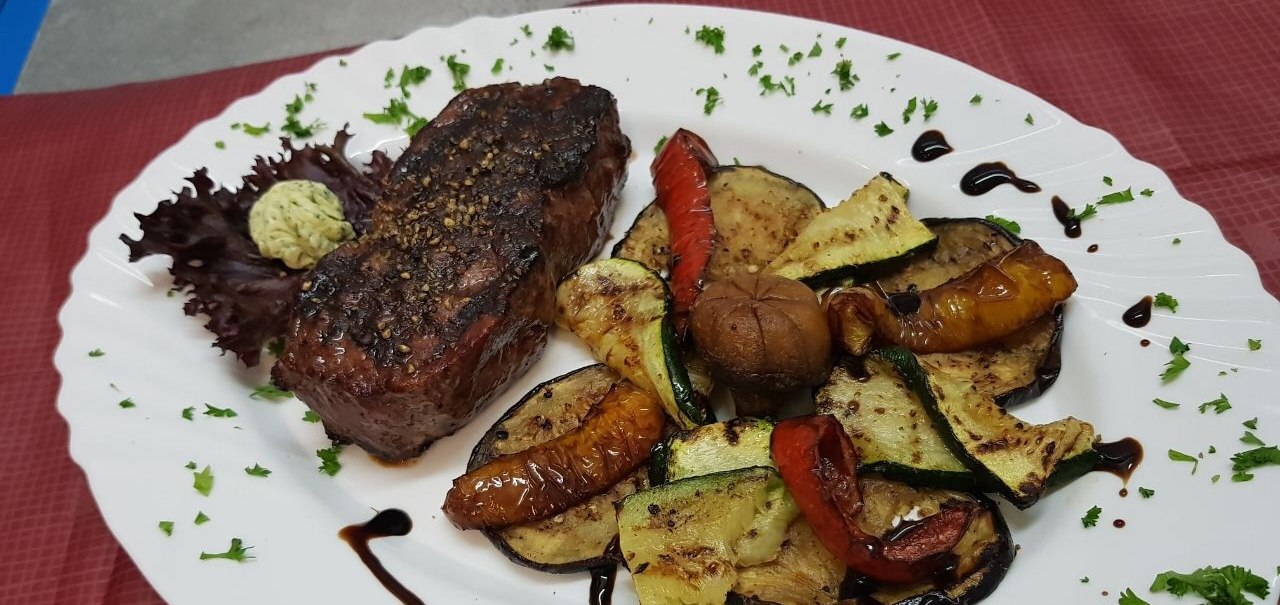 Gegrilltes Steak mit Gemüse und Kräuterbutter auf einem weißen Teller, dekoriert mit Balsamico und Petersilie, auf rotem Tischtuch serviert., © Restaurant Neckarau, P. Kotaidis