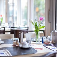 Ein Tisch in einem Café ist mit Geschirr, Besteck und einer Vase mit Tulpen gedeckt. Im Hintergrund sind Fenster und weitere Tische zu sehen., © TOMAS