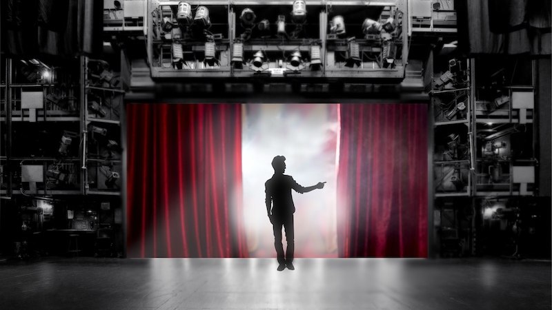 Ein Mann steht auf einer B&uuml;hne vor einem roten Vorhang, umgeben von B&uuml;hnentechnik. Der Hintergrund ist schwarz-wei&szlig;, der Vorhang rot., &copy; LIMA-Theater