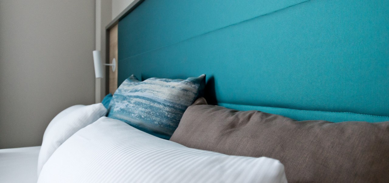 Nahaufnahme eines Bettes mit türkisfarbenem Kopfteil und Kissen in Weiß, Blau und Braun. Modernes, gemütliches Design., © Arthotel Ana / elaya