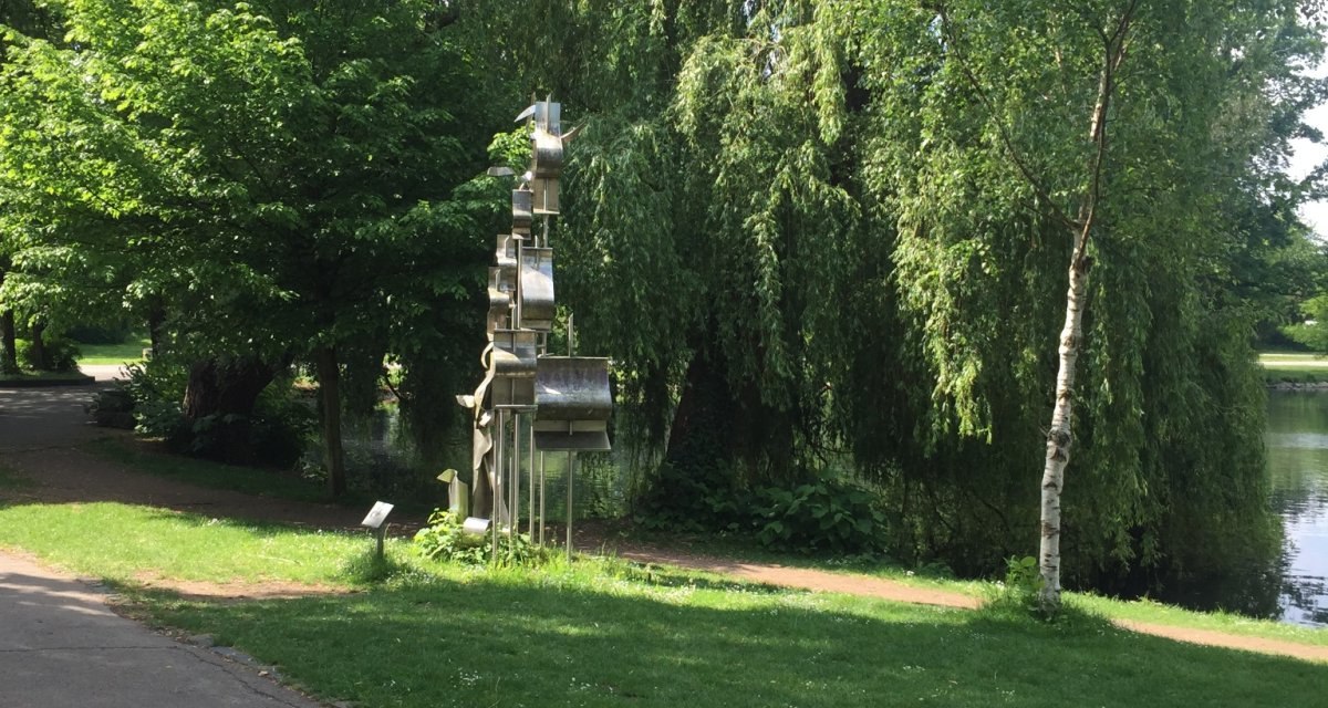 Eine Metallskulptur steht in einem grünen Park am Ufer eines Sees, umgeben von üppigen Bäumen und einem Gehweg., © www.pro-cycl.de Eine Metallskulptur steht in einem grünen Park am Ufer eines Sees, umgeben von üppigen Bäumen und einem Gehweg., © www.pro-cycl.de