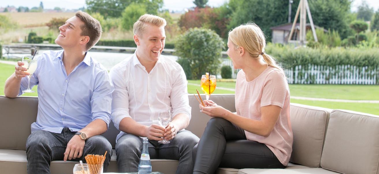 Drei Personen sitzen auf einer Terrasse, lachen und genießen Getränke. Im Hintergrund ist eine grüne Landschaft zu sehen., © ARAMIS Hotel & Freizeit GmbH
