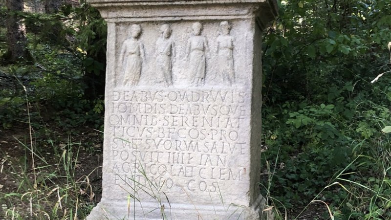 Ein steinernes Monument mit lateinischer Inschrift und vier eingravierten Figuren steht im Wald, umgeben von Bäumen und Gras., © Touristik und Marketing GmbH