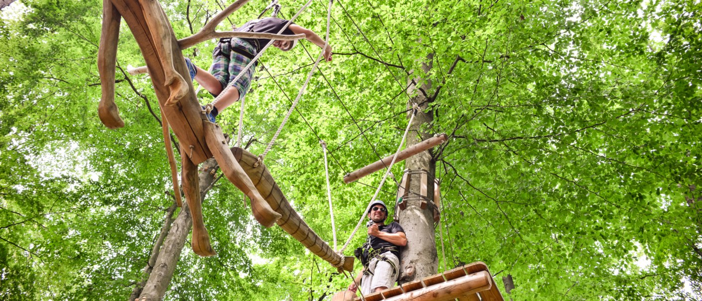 Zwei Personen klettern in einem Hochseilgarten. Eine Person balanciert auf einem Holzsteg, die andere steht auf einer Plattform. Umgeben von gr&uuml;nen Bl&auml;ttern., &copy; Kletterwald Plochingen