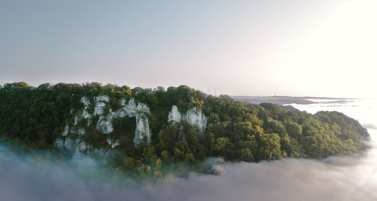 Ein bewaldeter Hügel mit markanten Felsformationen erhebt sich über Nebel. Im Hintergrund sind Windräder zu sehen, die in der Ferne stehen., © Landkreis Göppingen Ein bewaldeter Hügel mit markanten Felsformationen erhebt sich über Nebel. Im Hintergrund sind Windräder zu sehen, die in der Ferne stehen., © Landkreis Göppingen