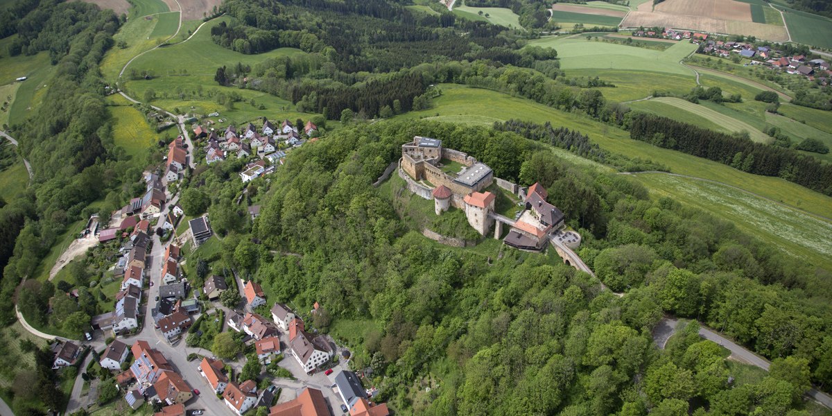 Luftaufnahme der Burgruine Rechberg in Schwäbisch Gmünd, umgeben von grünen Wäldern und einem kleinen Dorf. Felder und Wälder erstrecken sich im Hintergrund., © Burg Rechberg Luftaufnahme der Burgruine Rechberg in Schwäbisch Gmünd, umgeben von grünen Wäldern und einem kleinen Dorf. Felder und Wälder erstrecken sich im Hintergrund., © Burg Rechberg